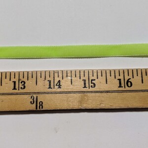 May include: A close-up of a bright green velvet ribbon measured against a wooden ruler. The ribbon is approximately 1 and 3/8 inches wide.