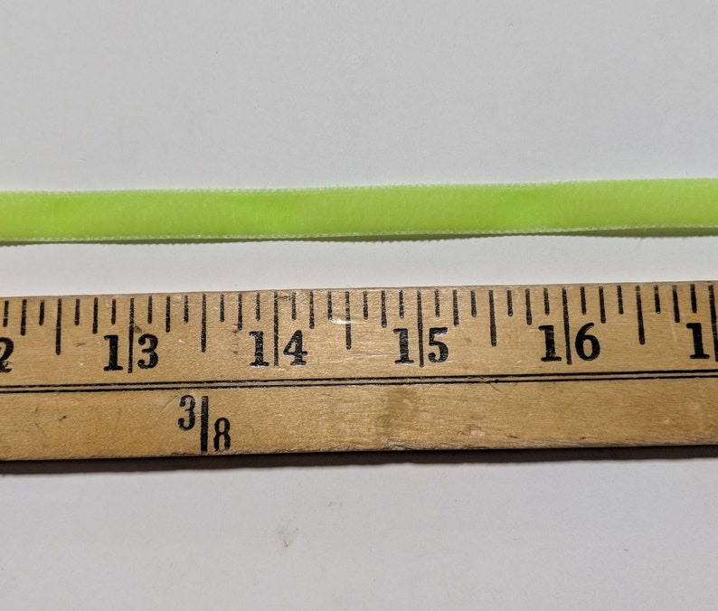 May include: A close-up of a bright green velvet ribbon measured against a wooden ruler. The ribbon is approximately 1 and 3/8 inches wide.