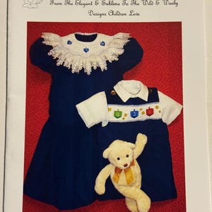 May include: A blue and white baby dress with a lace collar and a white embroidered design. A white and blue baby overall with a yellow and blue embroidered design. A teddy bear with a yellow bow is sitting on the dress. The image is titled "88 CHANUKAH".