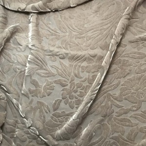 Op de afbeelding: Een beige fluwelen stof met een reliëf bloemenpatroon. De stof is zacht en luxueus, en het patroon is ingewikkeld en elegant.