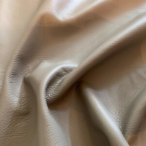 May include: A piece of taupe-colored leather with a natural grain texture. The material is draped to create soft folds and shadows, highlighting the smooth surface and the organic, irregular patterns of the hide's texture across the surface.