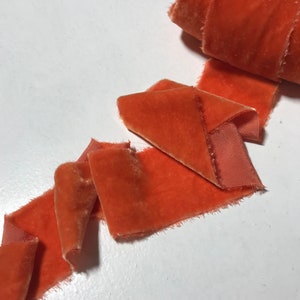 May include: A roll of burnt orange velvet ribbon with a frayed edge. The ribbon is folded and arranged on a white surface.