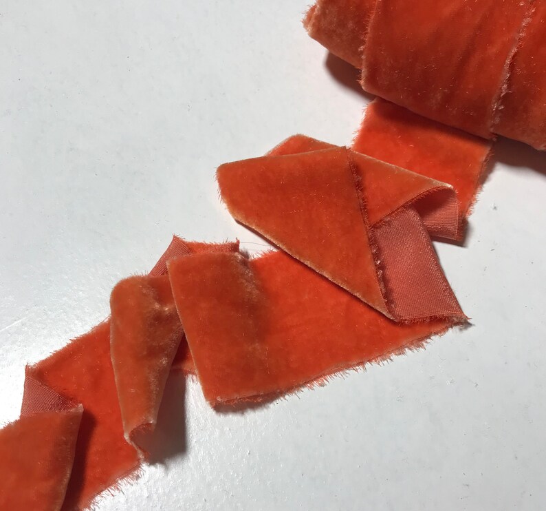 May include: A roll of burnt orange velvet ribbon with a frayed edge. The ribbon is folded and arranged on a white surface.