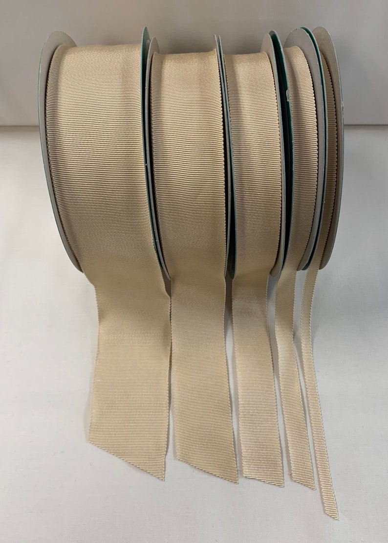 May include: Five spools of beige grosgrain ribbon in various widths. The ribbon is rolled and the ends are hanging down.