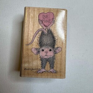 Puede incluir: Un sello de madera con una criatura caprichosa sosteniendo un corazón rosa con las palabras "BE MINE". La criatura tiene pelaje gris, orejas y pies rosas. El sello es rectangular y tiene el texto "©2004 Jareckie."