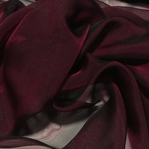 May include: Close-up of a flowing fabric in a deep burgundy colour. The sheer material has a slight sheen, with dark shadows creating depth and texture. The fabric is draped in soft folds, suggesting a lightweight, elegant textile.