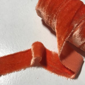 May include: A roll of burnt orange velvet ribbon with a frayed edge. The ribbon is unraveled and lying on a white surface.