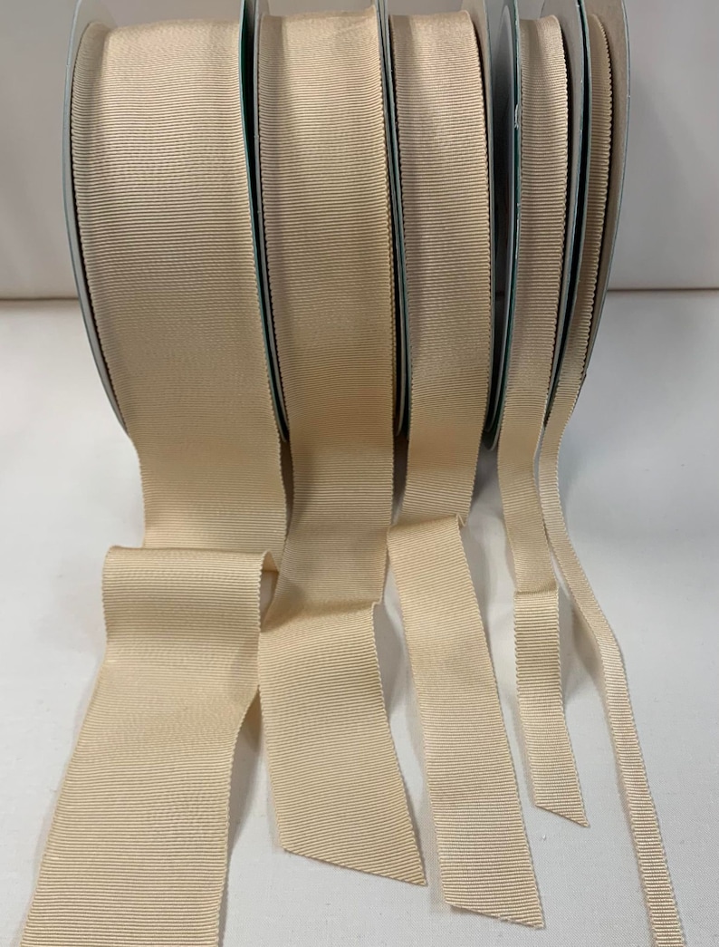 May include: Five spools of beige grosgrain ribbon in various widths. The ribbon is smooth and has a subtle sheen.