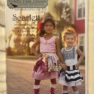 May include: A product image featuring two young girls modeling children's clothing. The image is a cover for a sewing pattern titled "Scarlett" by Violette Field Threads. The pattern includes an interchangeable tie-up over skirt, petti-skirt, and leg warmer set for sizes 2T-10 years.