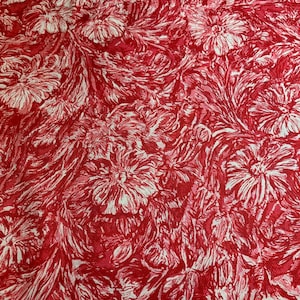 Pode incluir: Um tecido com um padrão denso e repetitivo de flores brancas e folhagem em espiral sobre um fundo vermelho vibrante. O design cria um motivo floral contínuo e intrincado que cobre toda a superfície do material têxtil.
