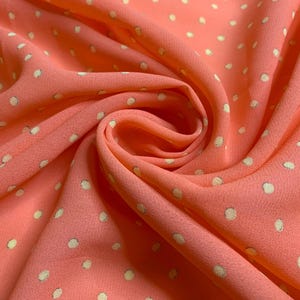May include: A coral-colored fabric featuring a repeating pattern of small, off-white polka dots. The material is gathered into a central swirl, showcasing the soft, draped texture of the textile with visible folds and light-catching highlights.