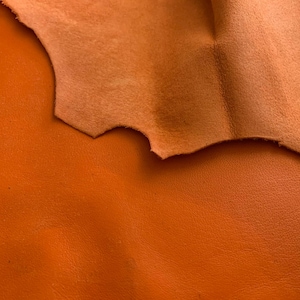 May include: A piece of vibrant orange leather with a smooth, matte finish on the front side and a lighter, textured suede-like surface visible on the folded edge. The material displays a consistent, saturated color throughout the visible area.