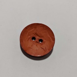 May include: A round, reddish-brown button with two small holes. The button has a textured surface and is likely made of wood or a similar material. This button is suitable for sewing projects.