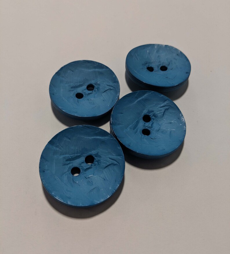 May include: Four blue plastic buttons with two holes each. The buttons have a textured surface and are arranged in a cluster.