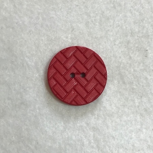 May include: A red plastic button with a woven pattern. The button has two holes.