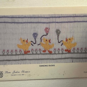 Könnte beinhalten: Ein Vintage-Stickmuster mit drei gelben Enten mit roten Füßen und Schleifen, die Luftballons halten. Die Enten tanzen auf einem hellblauen Hintergrund mit einem floralen Rand. Der Text "DANCING DUCKS" steht unter dem Bild.