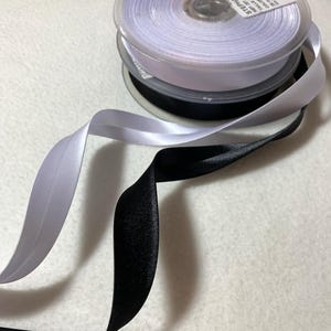 May include: Two spools of ribbon, one white and one black. The white ribbon is on top of the black ribbon. The white ribbon spool has a label that says "STEPHANIE 1/2" DOUBLE FACE SATIN 25 YARDS 100% POLYESTER MADE IN CHINA".