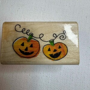 Może przedstawiać: Drewniana pieczątka z dwoma uśmiechniętymi dyniami w kolorze pomarańczowym i żółtym, połączonymi czarną winoroślą. Słowo "Cele" jest napisane nad dyniami czarną kursywą. Artykuł rzemieślniczy o tematyce Halloween.