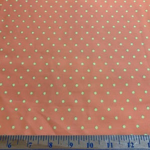 May include: A coral-colored fabric featuring a repeating pattern of small, light green polka dots. A measuring tape is positioned along the bottom edge, showing a scale in inches that spans from 14 to 28 inches across the width of the fabric.