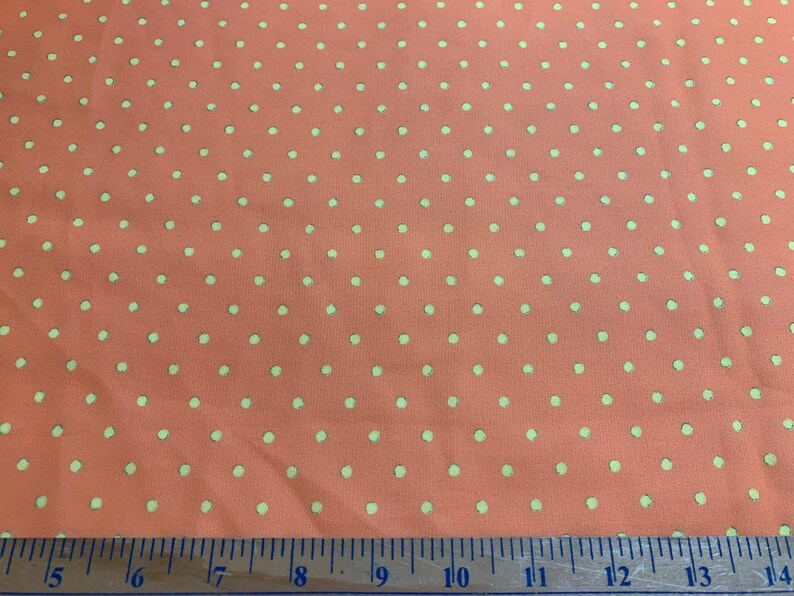May include: A coral-colored fabric featuring a repeating pattern of small, light green polka dots. A measuring tape is positioned along the bottom edge, showing a scale in inches that spans from 14 to 28 inches across the width of the fabric.