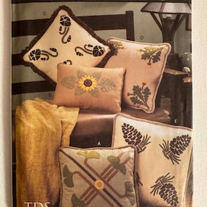 May include: A Vogue Patterns for Living sewing pattern envelope, number 7599, featuring illustrations of five decorative throw pillows with various nature-inspired applique designs, including sunflowers, pinecones, and oak leaves, with a gold fabric drape.