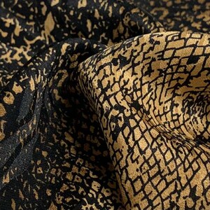 Puede incluir: Primer plano de una tela lujosa con un patrón texturizado. El material presenta una combinación de colores negro y dorado, con un diseño detallado que crea una apariencia rica y elegante. La tela está doblada, mostrando su profundidad y textura.