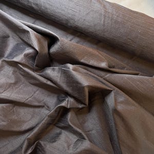 May include: A bolt of dark charcoal gray fabric with a subtle iridescent sheen and a visible slubbed texture. The material is draped in soft folds, highlighting the horizontal weave pattern and the deep, muted tones of the textile surface.