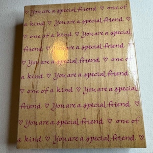Pode incluir: Um carimbo de madeira retangular com a frase "You are a special friend" repetida em rosa. A frase é intercalada com símbolos de coração. O carimbo é de cor castanho claro.