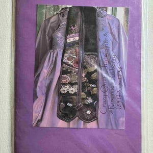 May include: A photograph of a purple garment with a decorative panel featuring floral and patterned fabric. The panel is dark with intricate details. The image is in a plastic sleeve.