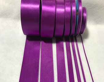 May include: Assortment of purple satin ribbon spools in various widths. The ribbon is arranged in a row, with the spools stacked on top of each other.
