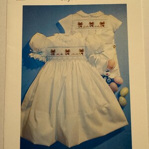 May include: A white baby dress with a brown embroidered bunny design. The dress has a ruffled collar and sleeves. The dress is paired with a white shirt with a brown embroidered bunny design. The shirt has a Peter Pan collar and short sleeves. The dress and shirt are laid out on a blue background with a stuffed bunny and Easter eggs.
