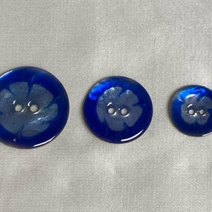以下が含まれることがあります： 白い花の模様が施された、虹色の青いボタンが3つ。ボタンにはそれぞれ2つの穴があります。