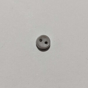 May include: A small, round, light gray button with two holes. The button is made of a smooth, glossy material and is suitable for sewing projects. The button is a neutral color and can be used on a variety of garments.