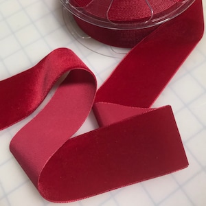 May include: A spool of rich burgundy velvet ribbon, partially unrolled, lies on a white grid surface. The ribbon's plush texture and deep color are highlighted, showcasing its luxurious quality. The ribbon is ideal for crafts and embellishments.