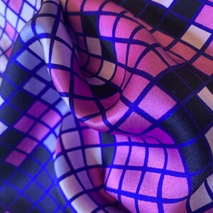Könnte beinhalten: Ein glatter Stoff mit einem sich wiederholenden Gittermuster aus Quadraten in Magenta-, Lila-, Lavendel- und Schwarztönen. Das Material ist in weichen Falten drapiert, was das geometrische Design und den seidenartigen Glanz der Oberfläche betont.