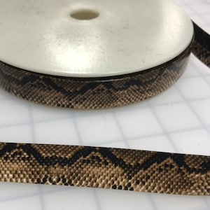 Puede incluir: Cinta de adorno con estampado de piel de serpiente marrón y negro. La cinta está enrollada en un carrete blanco.