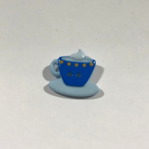 May include: A blue ceramic button with a white swirl on top, depicting a cup and saucer with yellow dots.