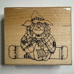 May include: A wooden stamp featuring a whimsical scarecrow design. The scarecrow is depicted with a straw hat, patched clothing, and a smiling face. The stamp is rectangular and has a light brown wood grain.