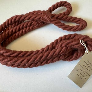 May include: A braided, rust-colored cotton tie back. The tie back is made of thick, twisted rope and has a loop at one end. A small tag is attached with the words "cotton traditions tie back" in English and French.