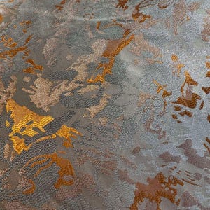 May include: A close-up of a silver and gold fabric with a textured, abstract pattern. The fabric has a subtle shimmer and a soft, luxurious feel.