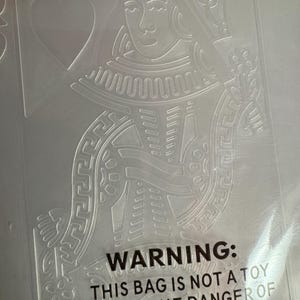 May include: A clear plastic embossing folder featuring a raised design of a playing card queen with a heart symbol. The text Cuttlebug and Provo Craft is printed at the top. A safety warning label regarding suffocation is printed at the bottom.