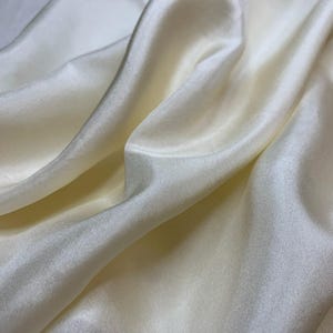 May include: A piece of off-white fabric with a smooth, lustrous surface and a satin-like sheen. The material is draped in soft, flowing folds, creating deep shadows and bright highlights that emphasize the reflective quality of the textile.