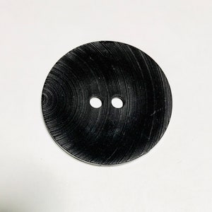 Peut inclure: Un bouton rond noir avec deux trous au centre. Le bouton a un motif en bois.