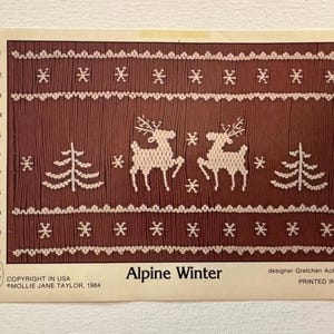 May include: A brown and white cross-stitch pattern titled "Alpine Winter." The design features white snowflakes, trees, and reindeer. The text "Alpine Winter" is displayed below the design, with the designer's name and copyright information.