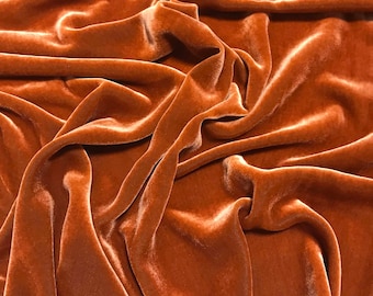 May include: Close-up of a luxurious, flowing fabric in a warm, earthy orange-brown hue. The material appears to be velvet, with soft folds and subtle highlights that catch the light, creating a rich, tactile texture. The fabric is draped, suggesting it's ready for use in apparel or home decor.