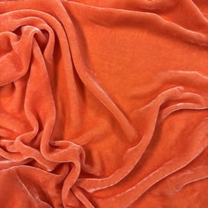 May include: Close-up of a vibrant orange velvet fabric with soft folds and a luxurious texture. The fabric has a subtle sheen, creating highlights and shadows across its surface. The material appears plush and inviting, perfect for apparel or home decor.