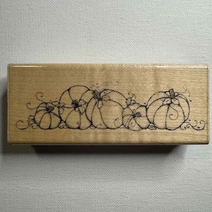 Puede incluir: Un sello de madera rectangular con un diseño de cinco calabazas de diferentes tamaños. Las calabazas están delineadas con tinta negra, con tallos detallados y zarcillos. El sello está hecho de madera clara.