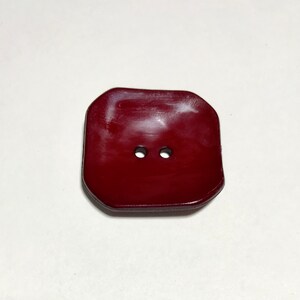 Può includere: Un bottone quadrato di colore rosso scuro con due fori, con una superficie leggermente rialzata e liscia.
