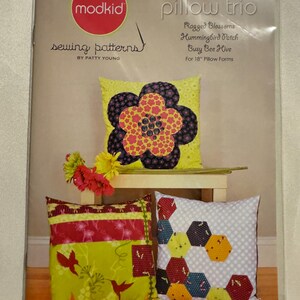 May include: A package of pillow sewing patterns by modkid. The package features three pillow designs: Ragged Blossoms, Hummingbird Patch, and Busy Bee Hive, designed for 18-inch pillow forms. The pillows have floral and geometric patterns in various colors.