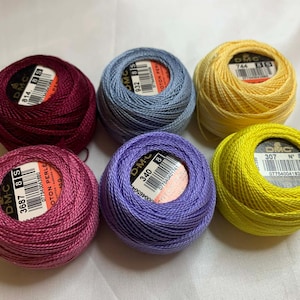 May include: Six spools of DMC Cotton Perle thread in various colors: burgundy, light blue, yellow, pink, purple, and chartreuse. Each spool has a label with a product code. The thread is tightly wound into a ball.